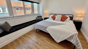 Een slaapkamer met een groot raam, een bed met veel kussens, een nachtkastje met een lamp en een plafondventilator.