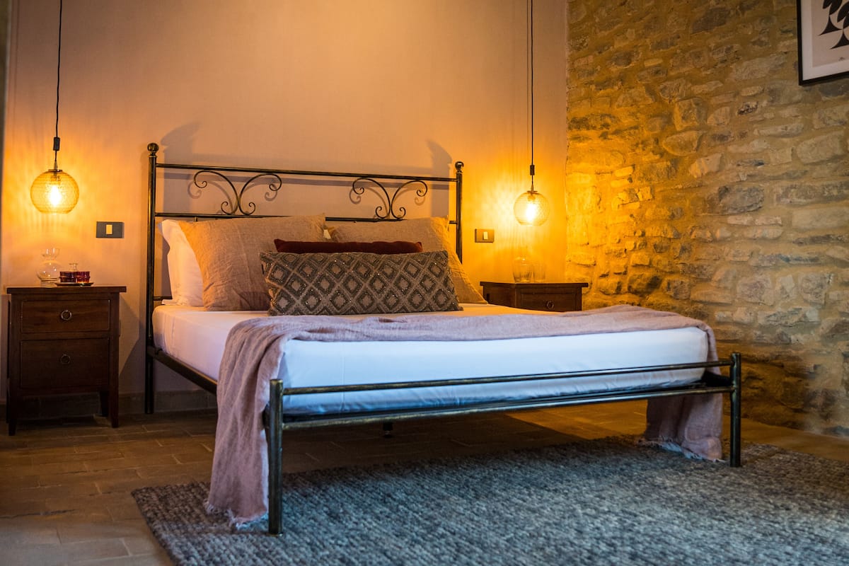 Una cama con estructura metálica, dos mesitas de noche, una pared de piedra y dos lámparas de pared.