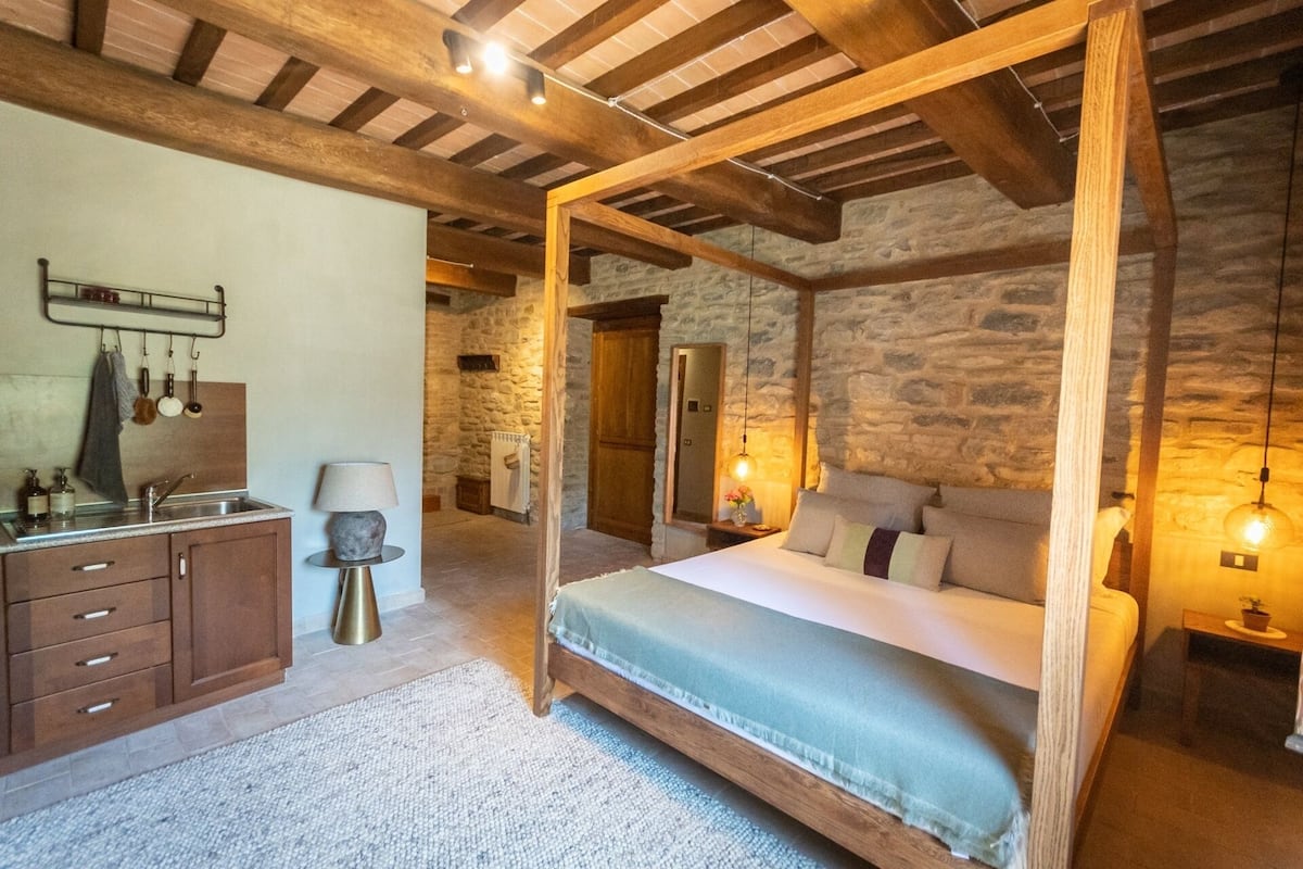 Un dormitorio rústico con cama con dosel, paredes de piedra, vigas de madera, chimenea de piedra y una pequeña área de cocina con lavamanos y armarios.