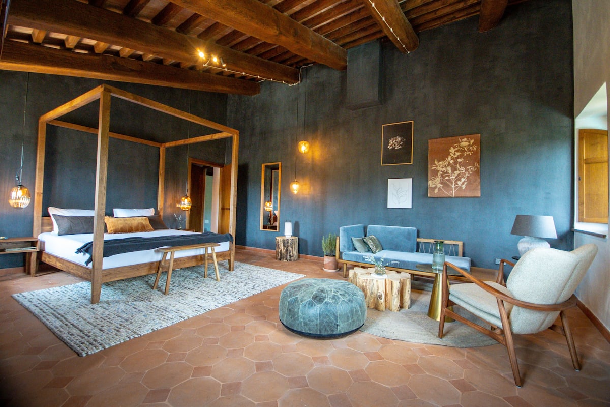 Un dormitorio con cama con dosel, un sofá, una silla y un reposapiés redondo.