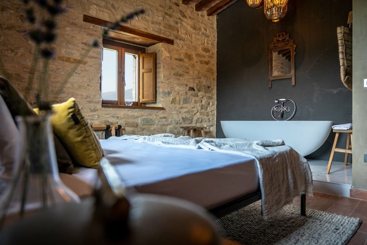 Un dormitorio con pared de piedra, una cama grande, cabecera de madera, una ventana, una bañera y un espejo en la pared.