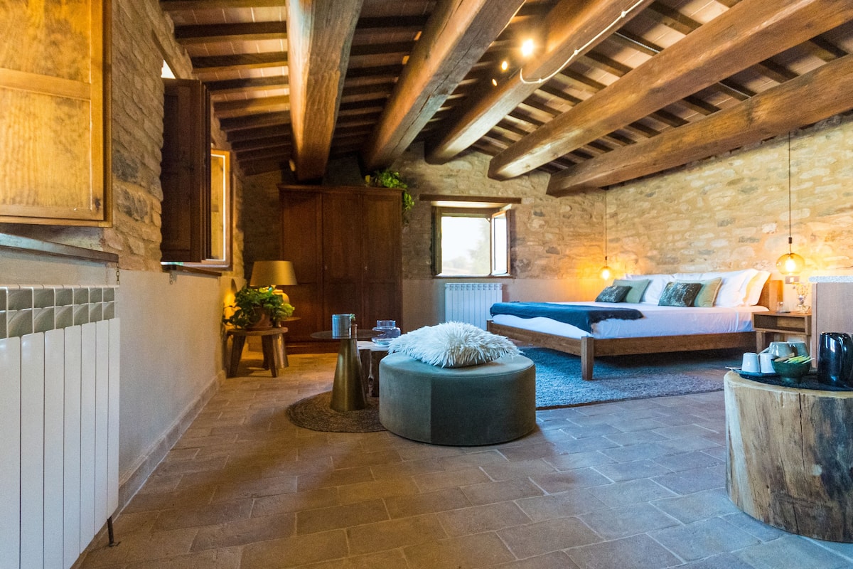 Una habitación acogedora con pared de piedra, vigas de madera, una cama, un sofá y un reposapiés redondo.