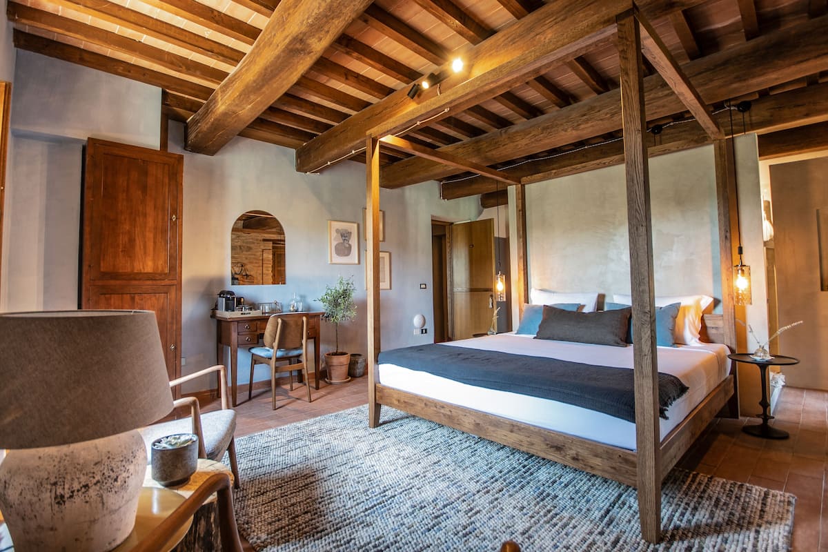 Un dormitorio con cama con dosel, vigas de madera en el techo, un escritorio con silla y una mesita de noche con lámpara.