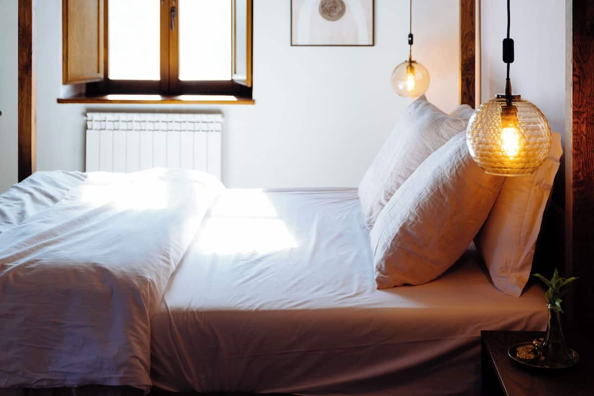 Un dormitorio con un ventanal grande, un radiador, una cama con ropa de cama blanca y dos lámparas colgantes.