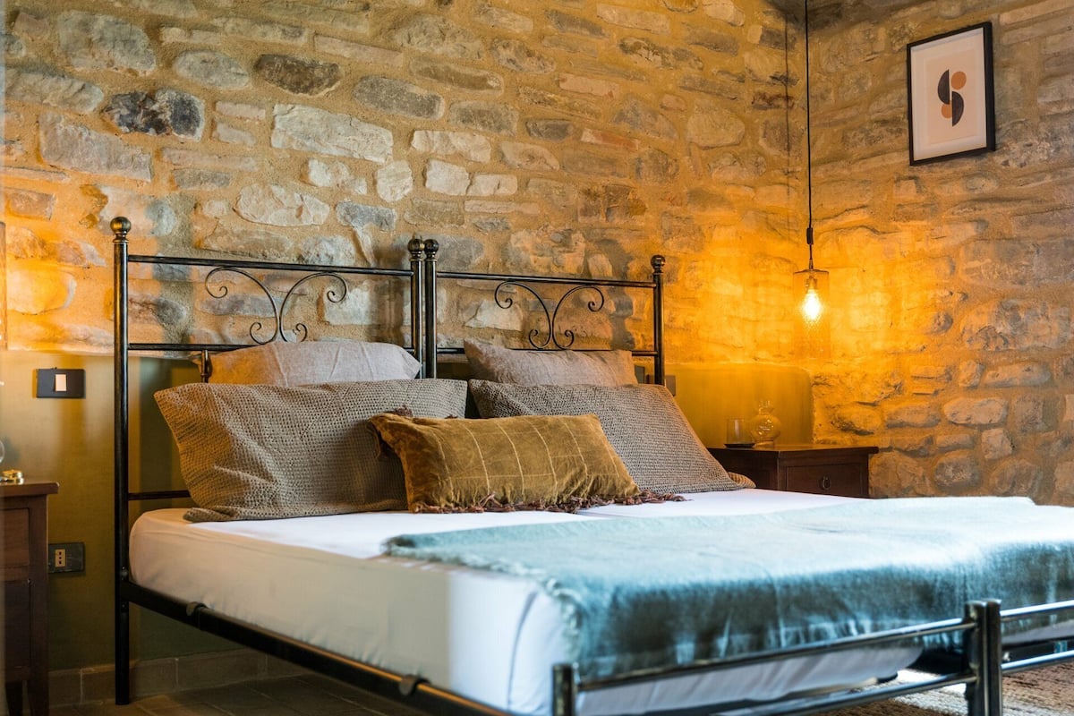 Una cama con estructura metálica, varios almohadones y una colcha en una habitación con paredes de piedra y una lámpara colgante.