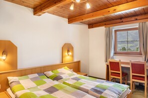 A room with a wooden ceiling, a bed with a checkered bedspread, a desk with a chair, and a window with curtains.