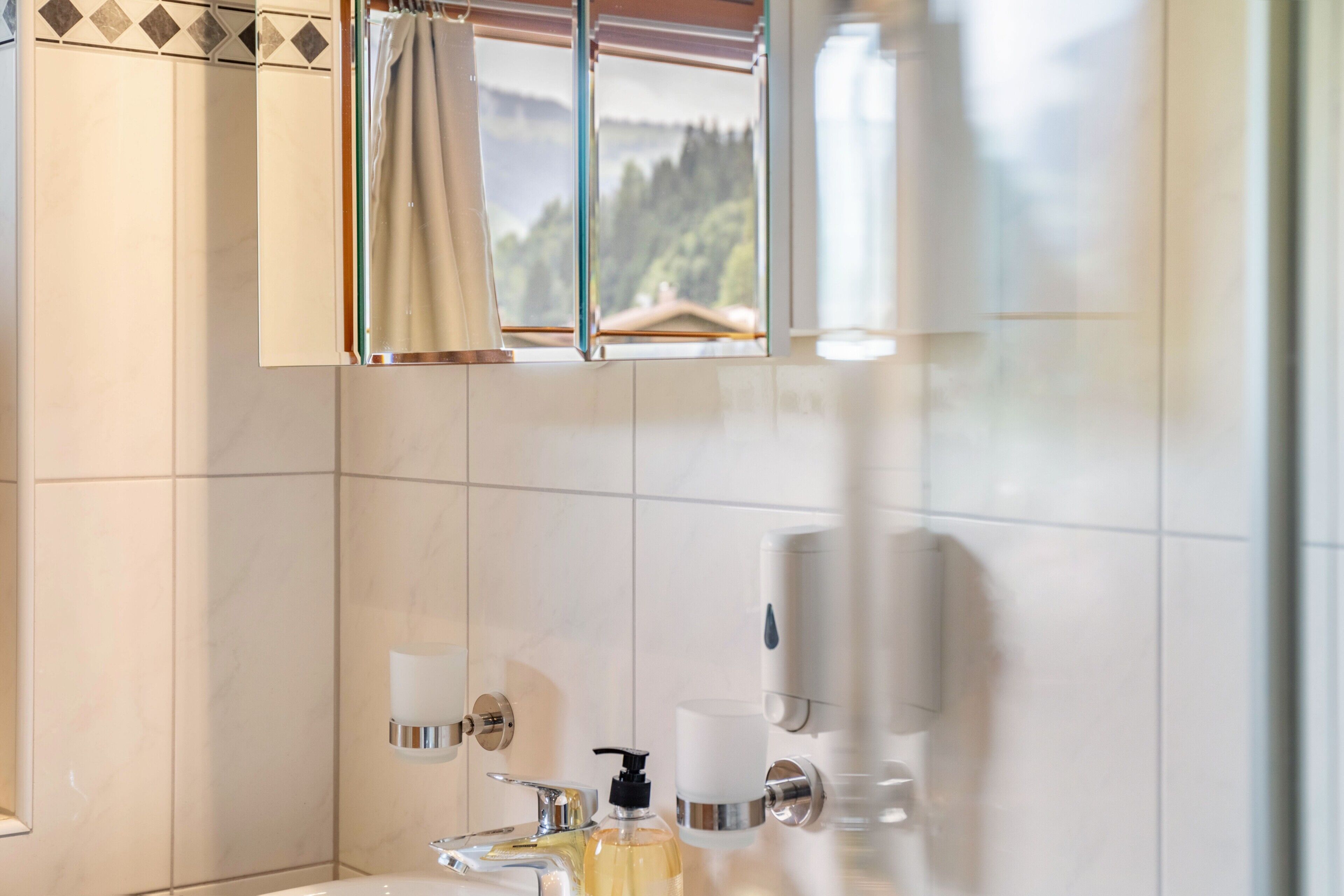 Une salle de bain avec une fenêtre, un miroir, un lavabo, un distributeur de savon et un porte-serviettes.