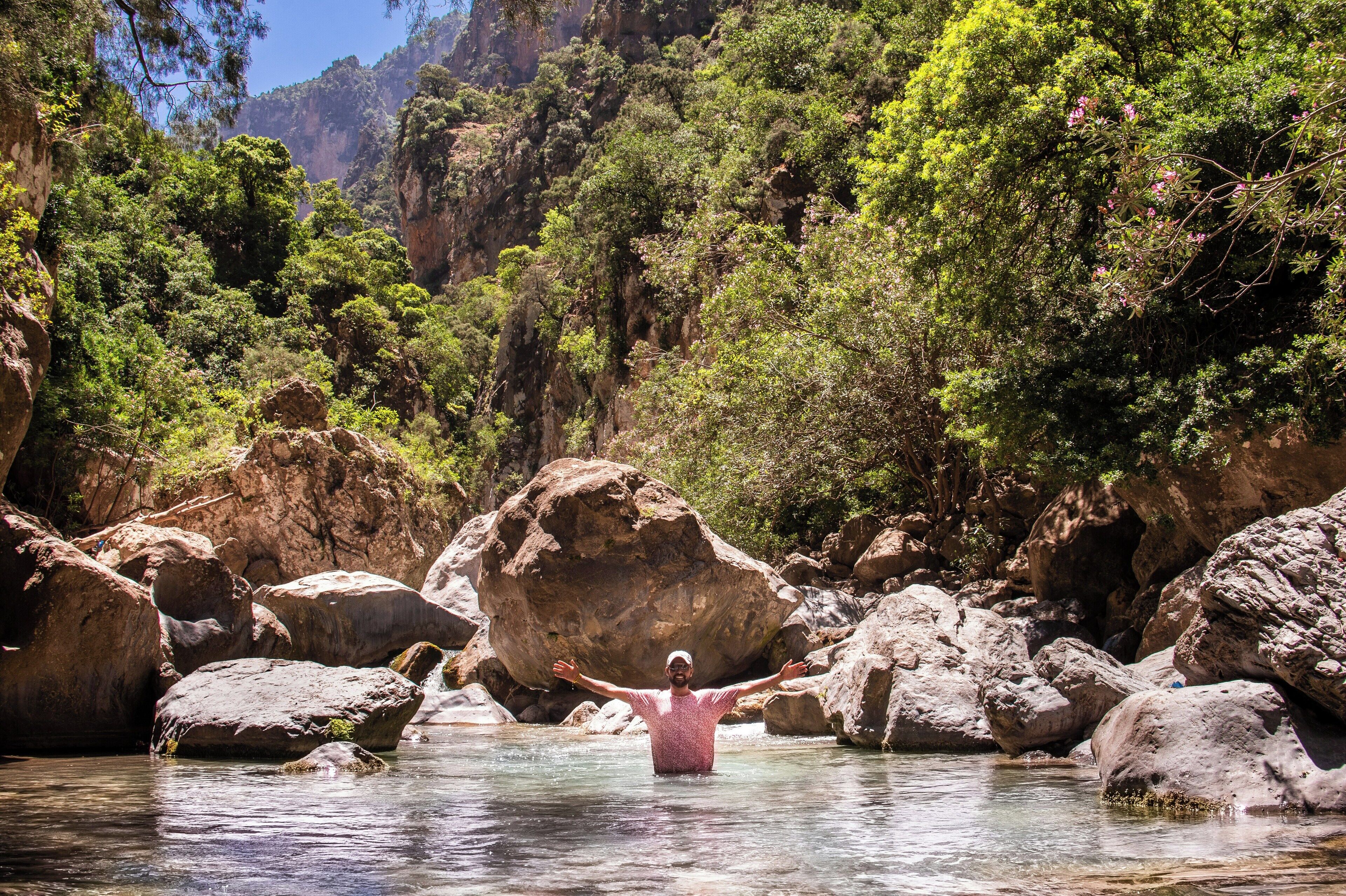 Une personne se tient dans une rivière, entourée de gros rochers et d’une végétation luxuriante.