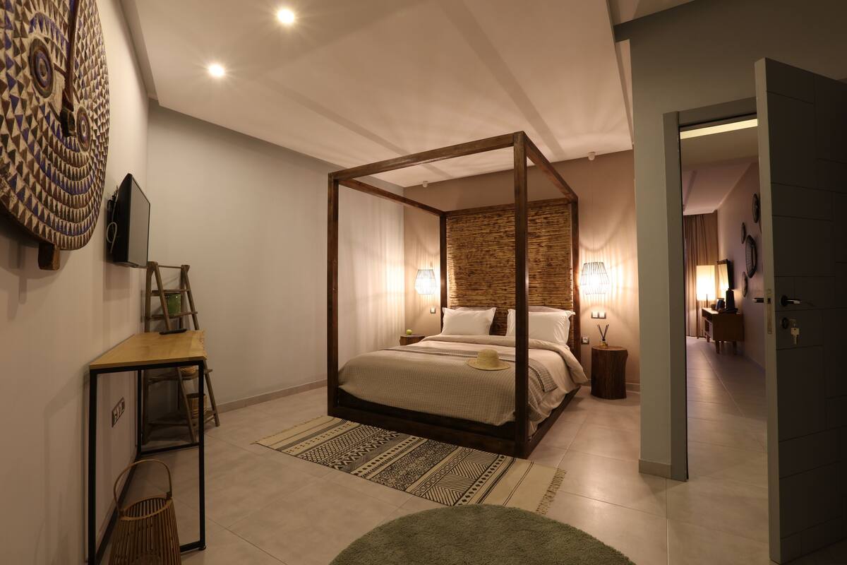 A modern bedroom with a four-poster bed, a wooden desk, and a large decorative wall piece.