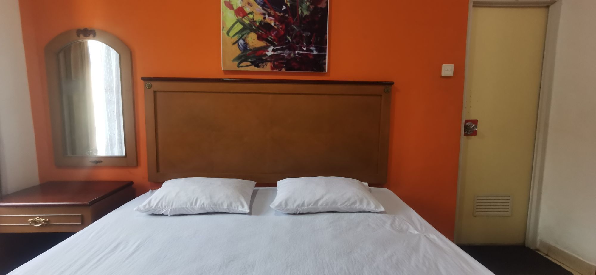 A hotel room with a large bed, two pillows, a wooden headboard, a mirror, a dresser, and a painting on the wall.