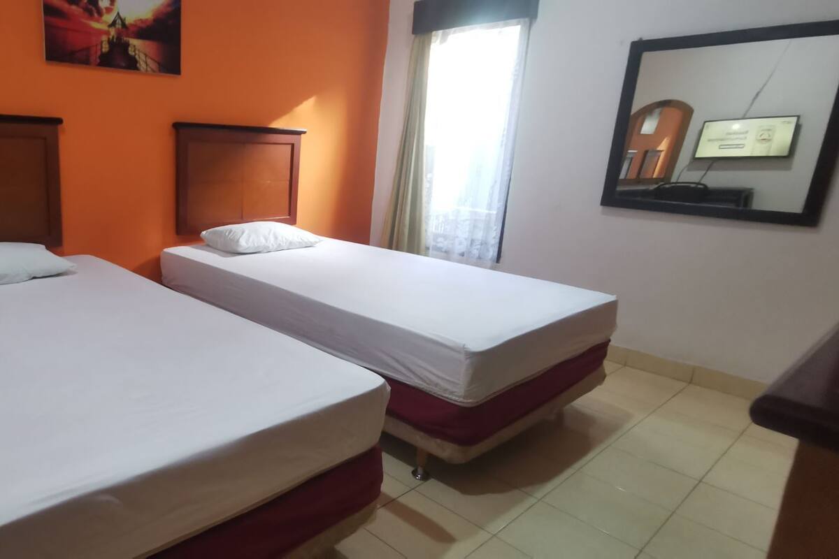 A room with two single beds, a wooden headboard, a window with curtains, and a wall-mounted TV.