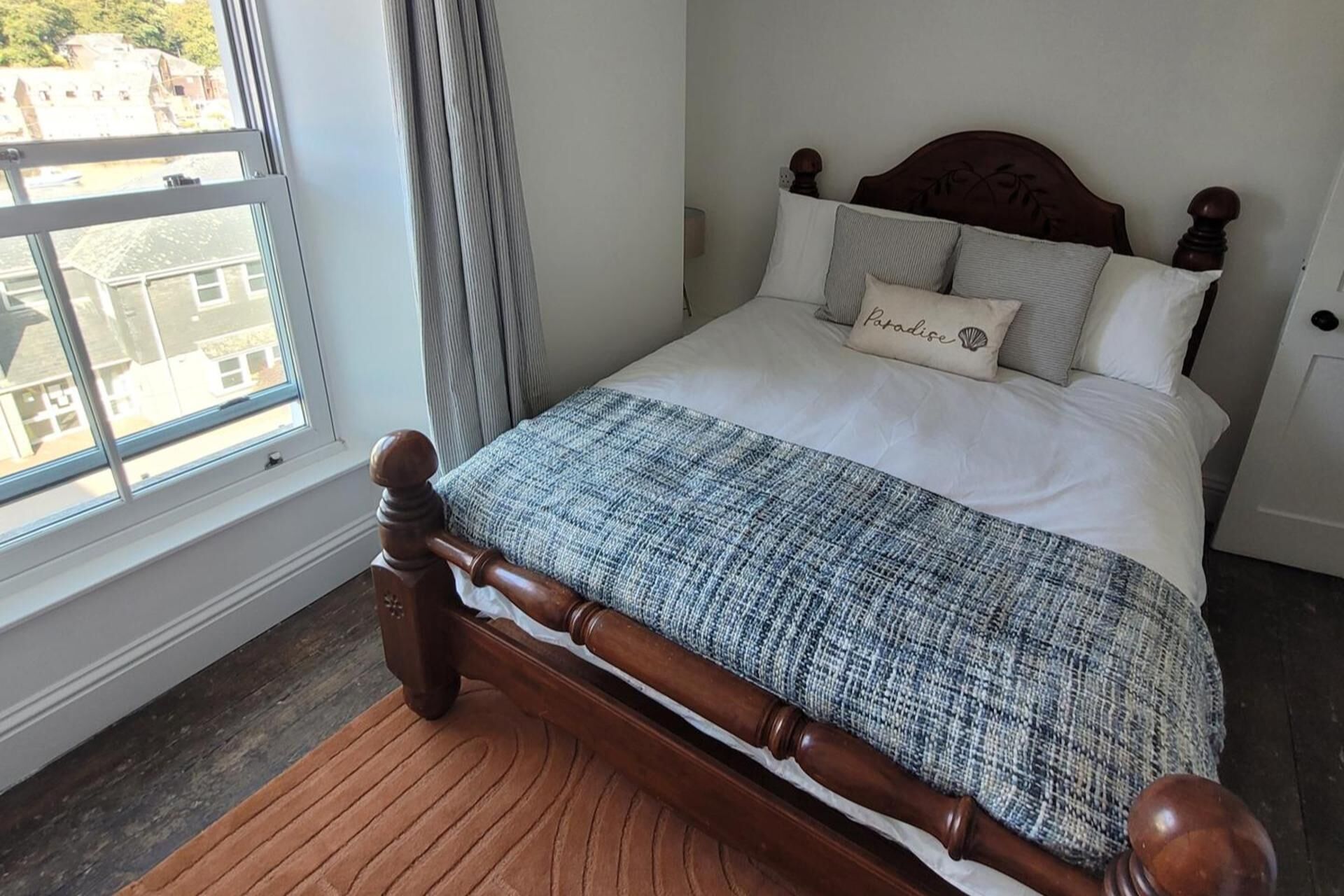 Un lit bien fait, avec une tête de lit en bois, une fenêtre donnant sur des immeubles et un plancher en bois.