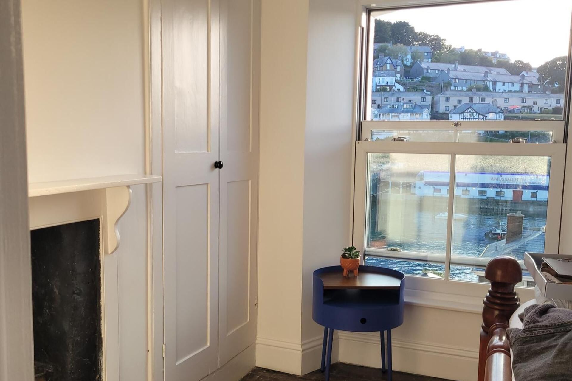 A room with a white door, a small blue table with a plant, and a view of a town through a window.