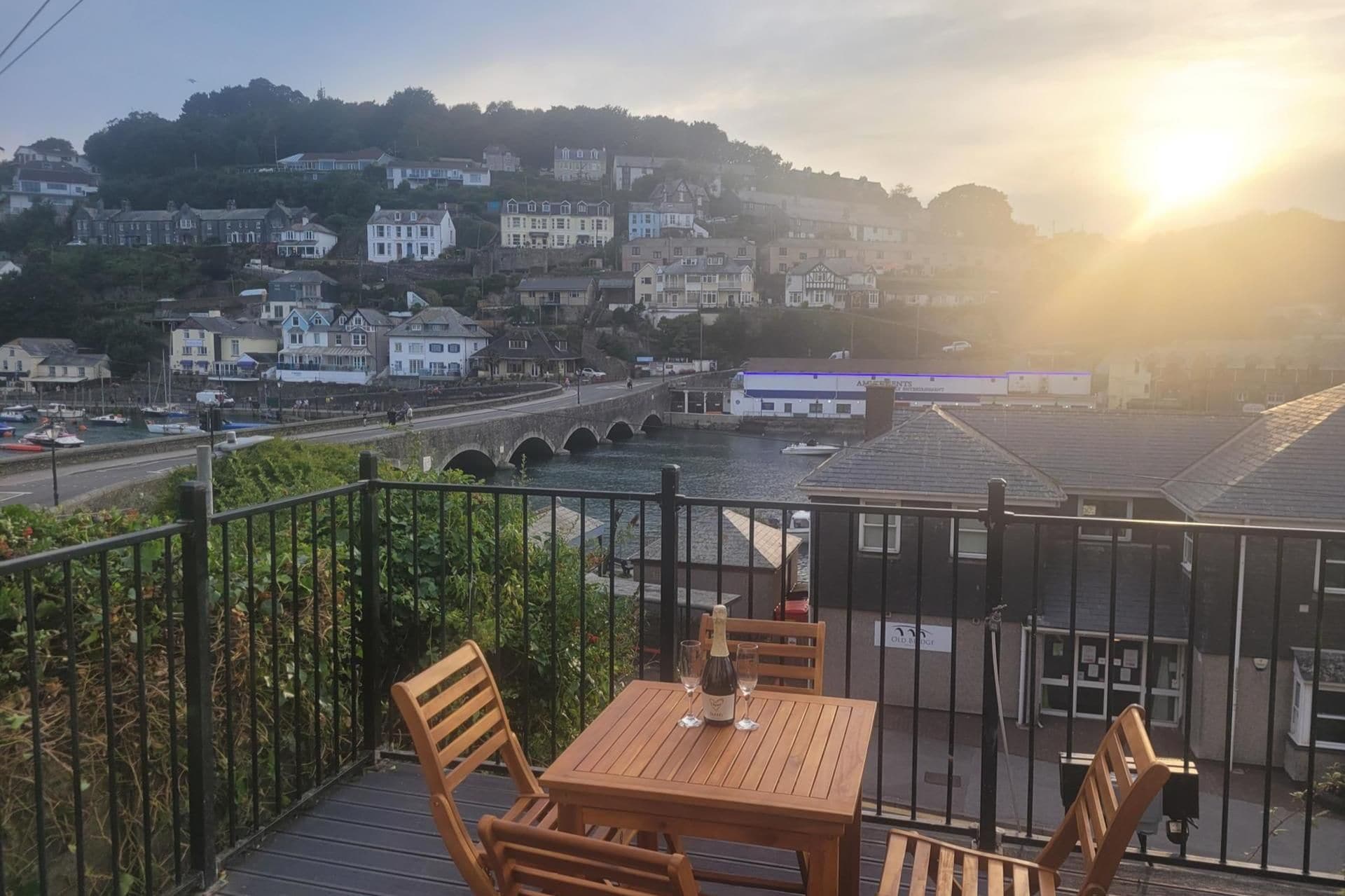 A balcony with a table and chairs overlooking a river, a bridge, and a hillside of buildings.