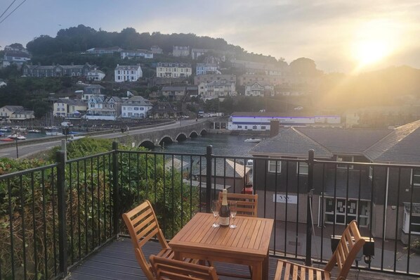 A balcony with a table and chairs overlooking a river, a bridge, and a hillside of buildings. - Looe Cottage - Seaside Views, log Fire, Sleeps 4 (Looe)