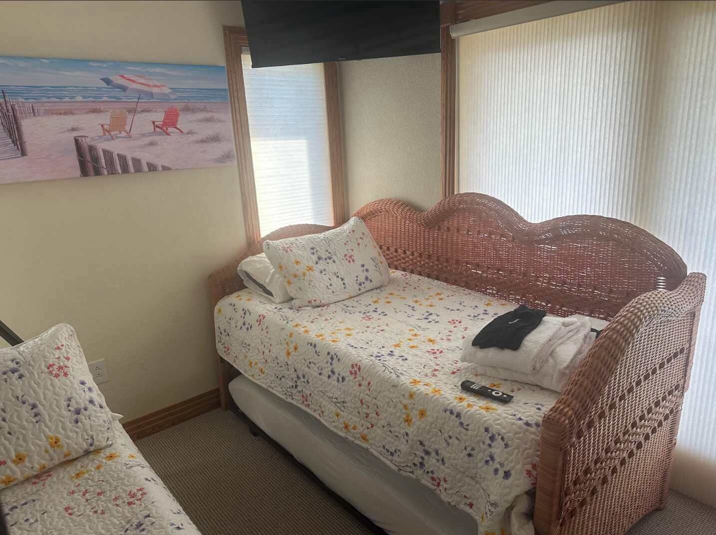 Une petite pièce agréable, avec un lit simple et une chaise, un tableau représentant une scène de plage, et une fenêtre avec des stores.