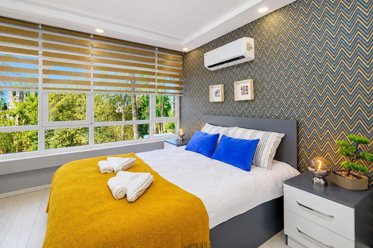 A modern hotel room with a large bed, a bedside table, a nightstand, a window with blinds, an air conditioning unit, and a decorative wall pattern.