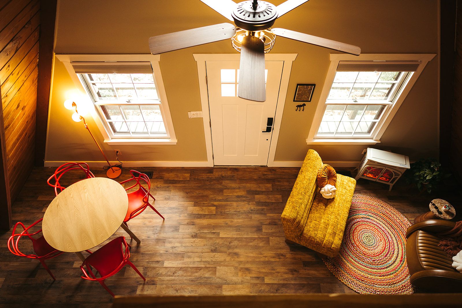 A room with a wooden floor, a ceiling fan, a dining table with red chairs, and a sofa with a colorful rug.