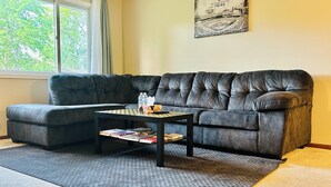 A living room with a sectional sofa, a coffee table with books and a water bottle, a rug, and a framed picture on the wall.