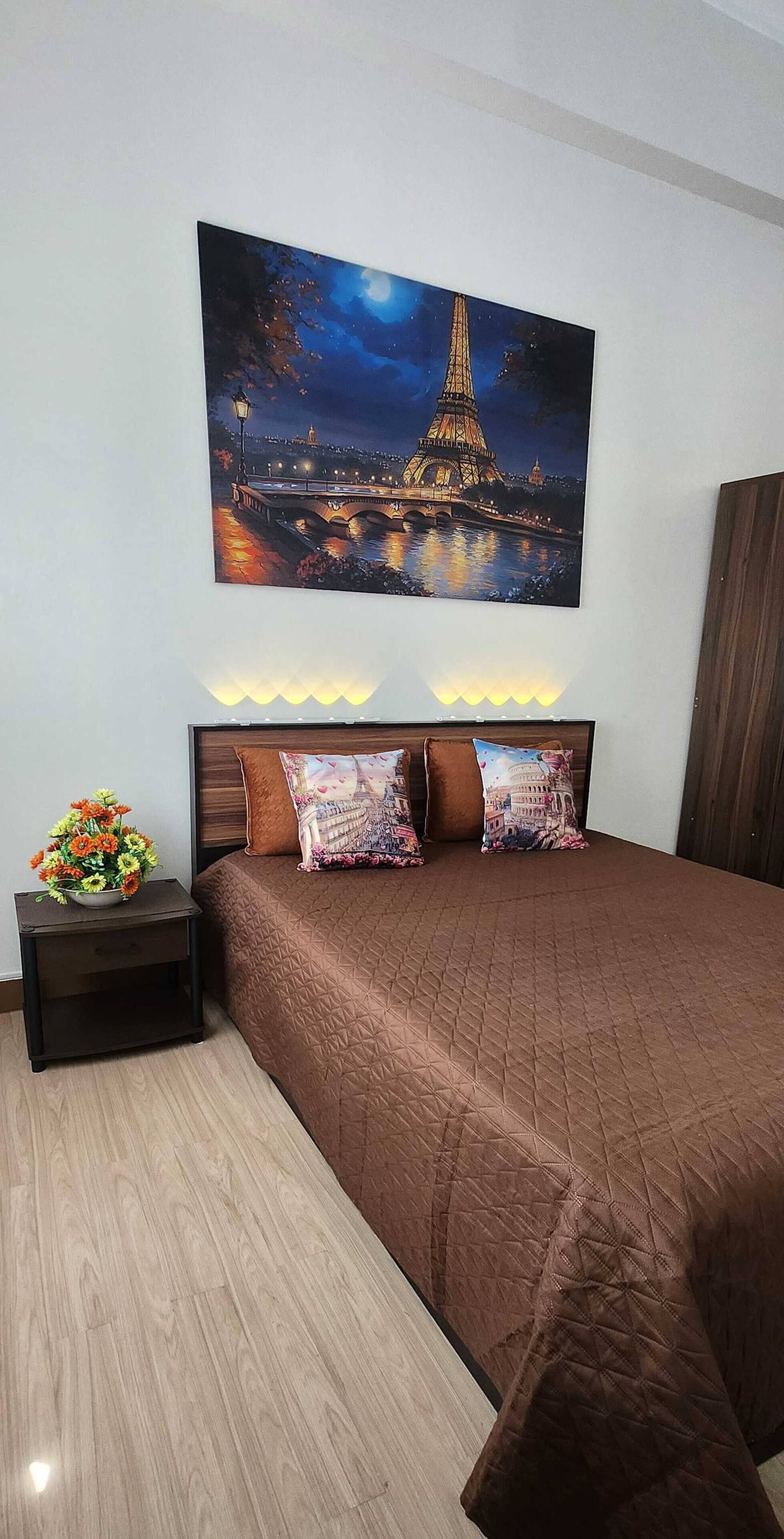Un dormitorio con una cama grande, vista nocturna de la Torre Eiffel, una mesita de noche con flores y suelo de madera.