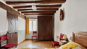 Una habitación con vigas de madera a la vista, una cama con colcha, un armario de madera, una silla y un escritorio con lámpara.