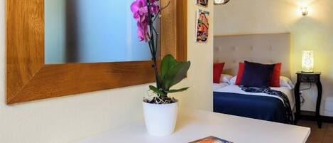 A bedroom with a bed, a bedside table with a book, a potted plant, and a mirror on the wall.