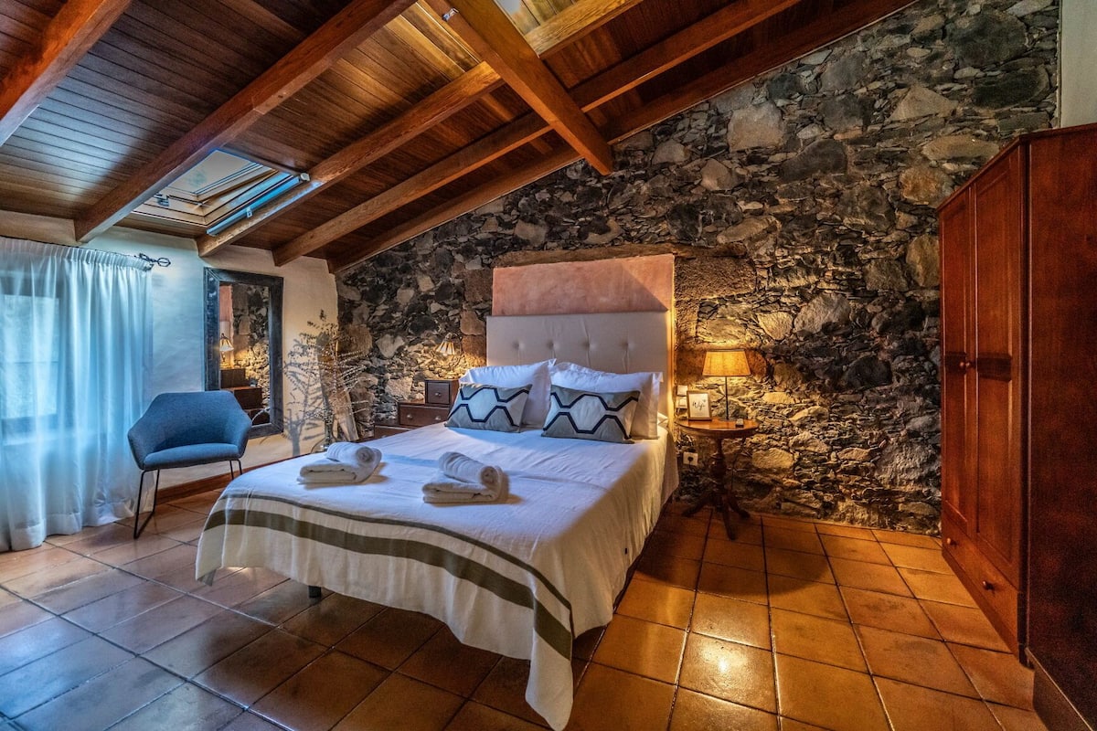 A bedroom with a stone wall, wooden ceiling beams, a bed with pillows, a chair, and a nightstand with a lamp.
