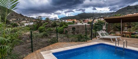 A swimming pool with lounge chairs and a wooden deck, surrounded by a fence and greenery, with a view of a town and hills in the background.