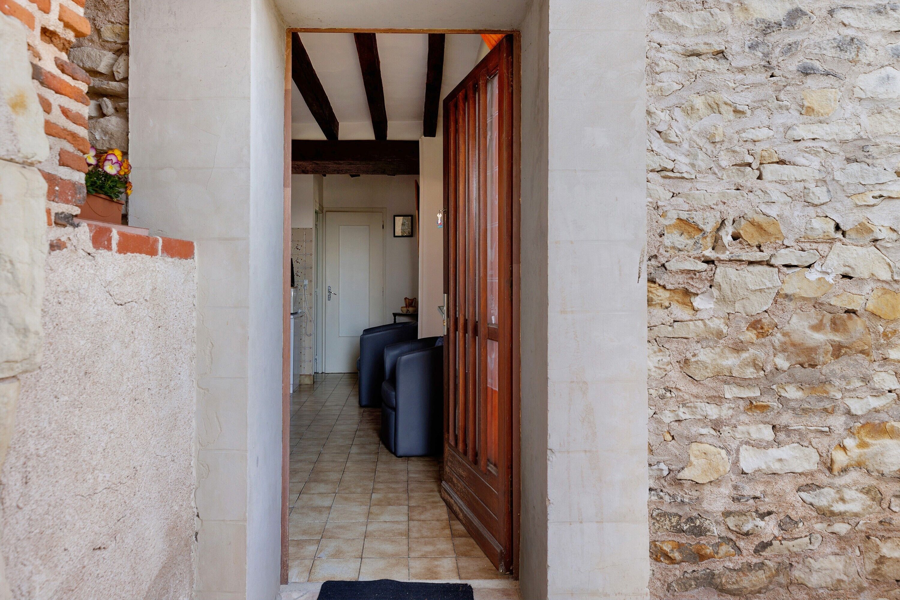 A narrow hallway with a stone wall on one side and a wooden door on the other. There are two black chairs in the distance.