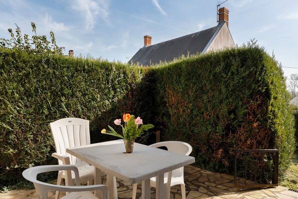 A patio with a table and chairs, a vase with flowers, and a hedge in the background.