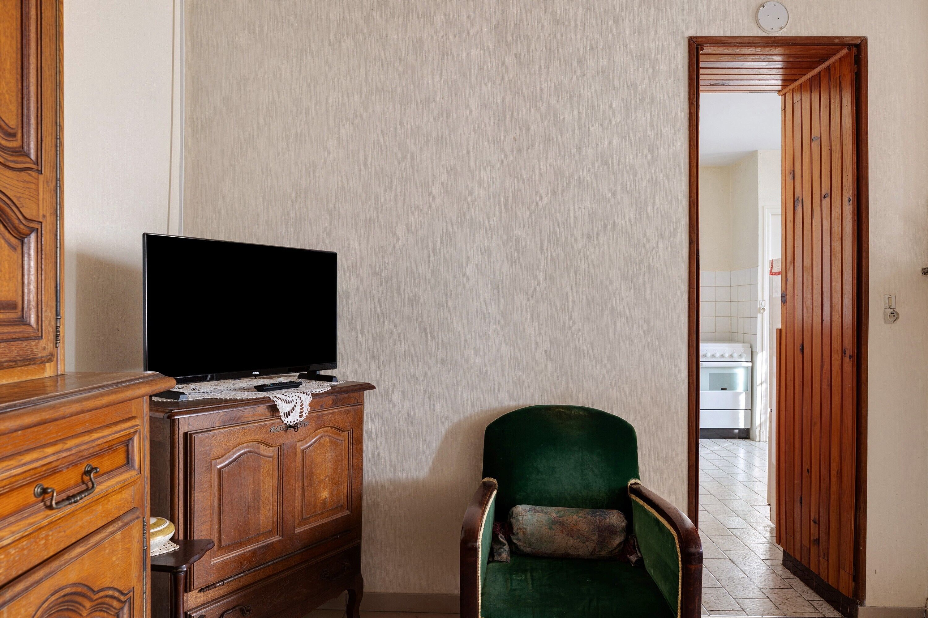 Une pièce avec une commode en bois, un fauteuil vert, un téléviseur posé sur le dessus, et une cuisine visible à travers une porte ouverte.