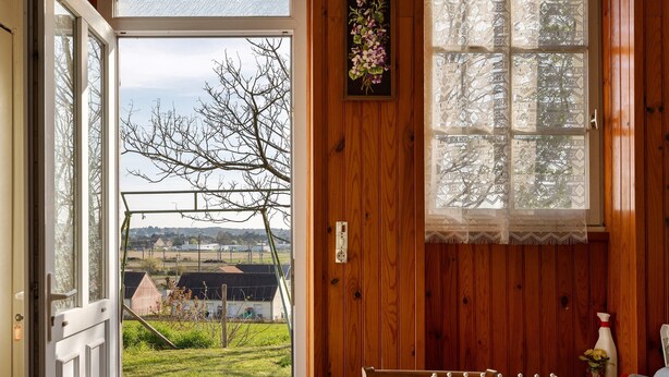 Ein Zimmer mit Blick auf eine ländliche Landschaft, eine Holztür, ein Tisch mit einer Tischdecke mit Blumenmuster und ein Fenster mit gemusterten Vorhängen.