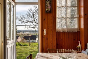 A room with a view of a rural landscape, a wooden door, a table with a floral tablecloth, and a window with patterned curtains.
