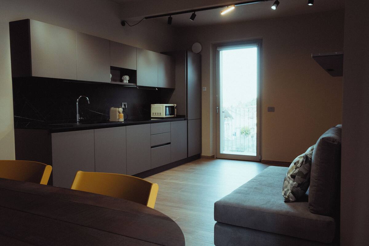 A modern kitchen with grey cabinets, a black countertop, and a microwave. There is a dining area with a table and chairs, and a grey sofa with a cushion.