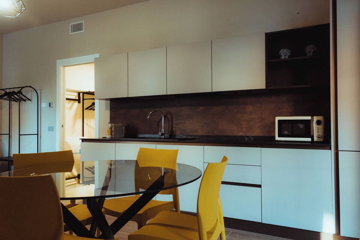 A modern kitchen with a glass dining table, yellow chairs, white cabinets, and a microwave.