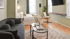 Ein modernes Wohnzimmer mit einer Couch, einem Glastisch, einem Bett mit weißer Bettdecke, einem an der Wand befestigten Fernseher und einem Fenster mit Vorhängen.