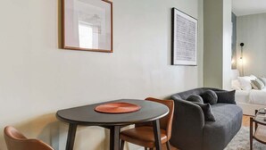 Ein modernes Wohnzimmer mit einem runden Esstisch, zwei braunen Ledersesseln, einem grauen Sofa und einem kleinen Couchtisch. An der Wand hängen zwei gerahmte Bilder, und eine Deckenleuchte ist sichtbar.