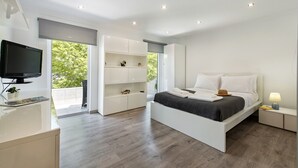 A modern bedroom with a large bed, built-in shelving, a flat-screen TV, and a balcony with a view of greenery. - Casa dos Quatro in Faro (Faro)