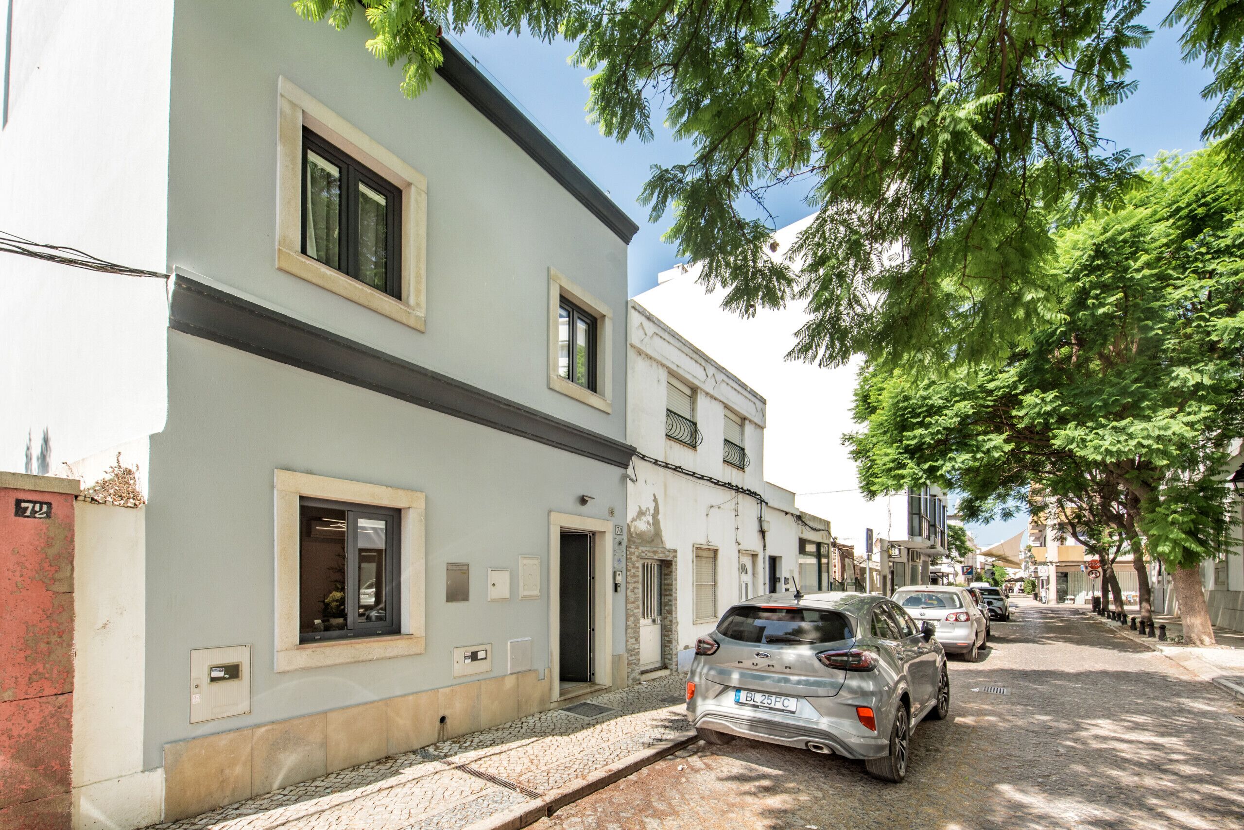 A street view with a row of parked cars, a building with a light facade, and trees lining the street.