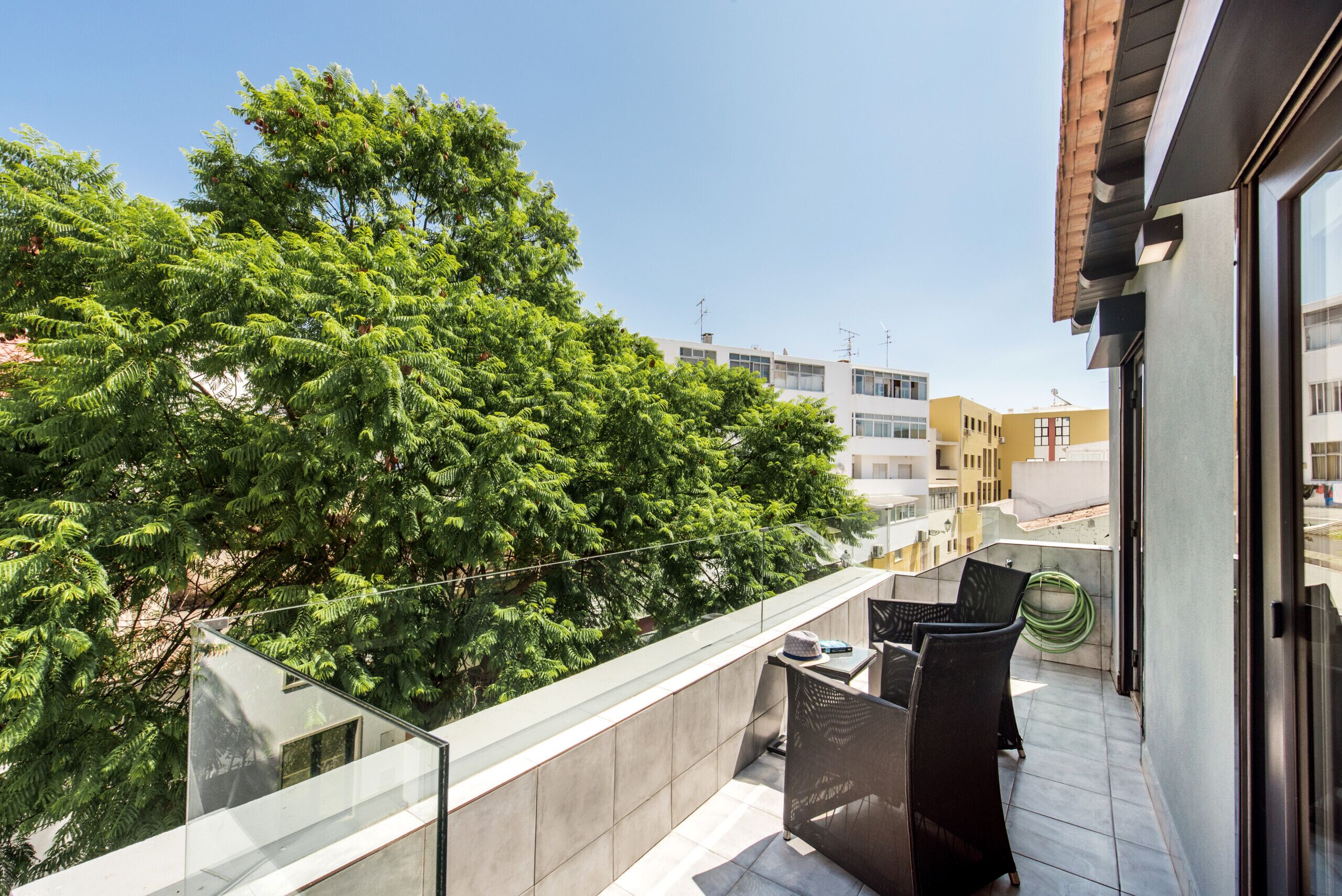 Un balcon doté d’une balustrade en verre, de mobilier d’extérieur et offrant une vue sur les bâtiments et les arbres environnants.