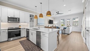 Une cuisine moderne avec des meubles blancs, des appareils en acier inoxydable et un îlot central. On y trouve des luminaires suspendus, un ventilateur de plafond et une vue sur l’extérieur grâce à de grandes fenêtres.