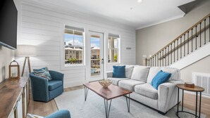 Un salon spacieux avec un canapé gris, un fauteuil bleu et une table basse en bois. On aperçoit un escalier au fond.
