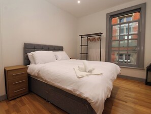 Un lit bien fait, avec du linge de lit et des serviettes blanches, une table de chevet en bois dotée de tiroirs, une tête de lit rembourrée de couleur grise et une fenêtre donnant sur un bâtiment en briques.