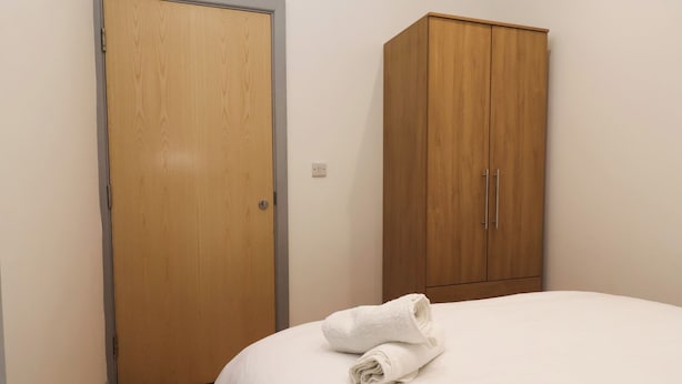 A bedroom with a wooden wardrobe, a bed with white bedding, and a rolled white towel on the bed.