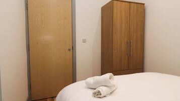 A bedroom with a wooden wardrobe, a bed with white bedding, and a rolled white towel on the bed.