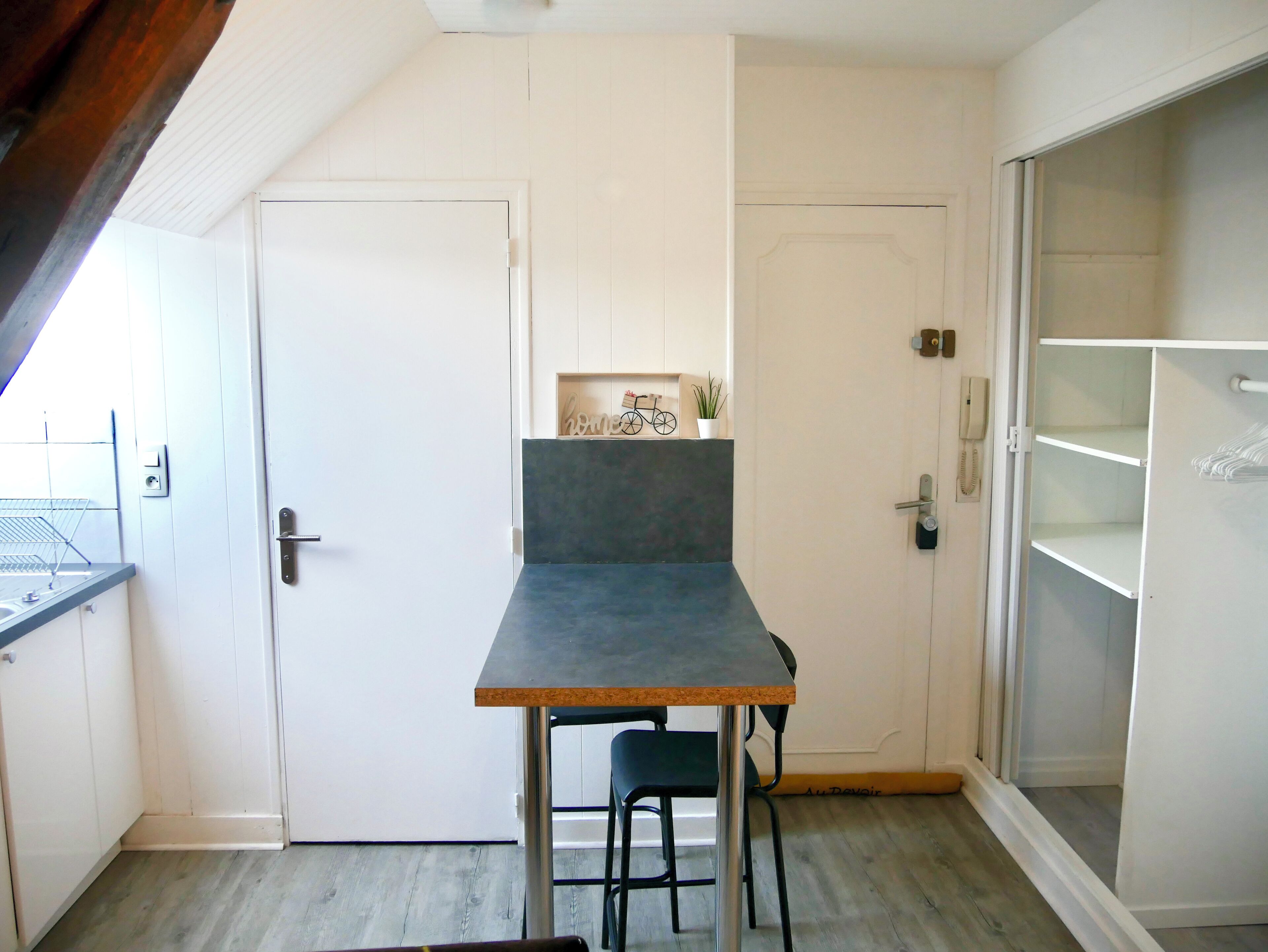 Een compacte keuken met een kleine eettafel en stoel, een witte deur en een inbouwkast.