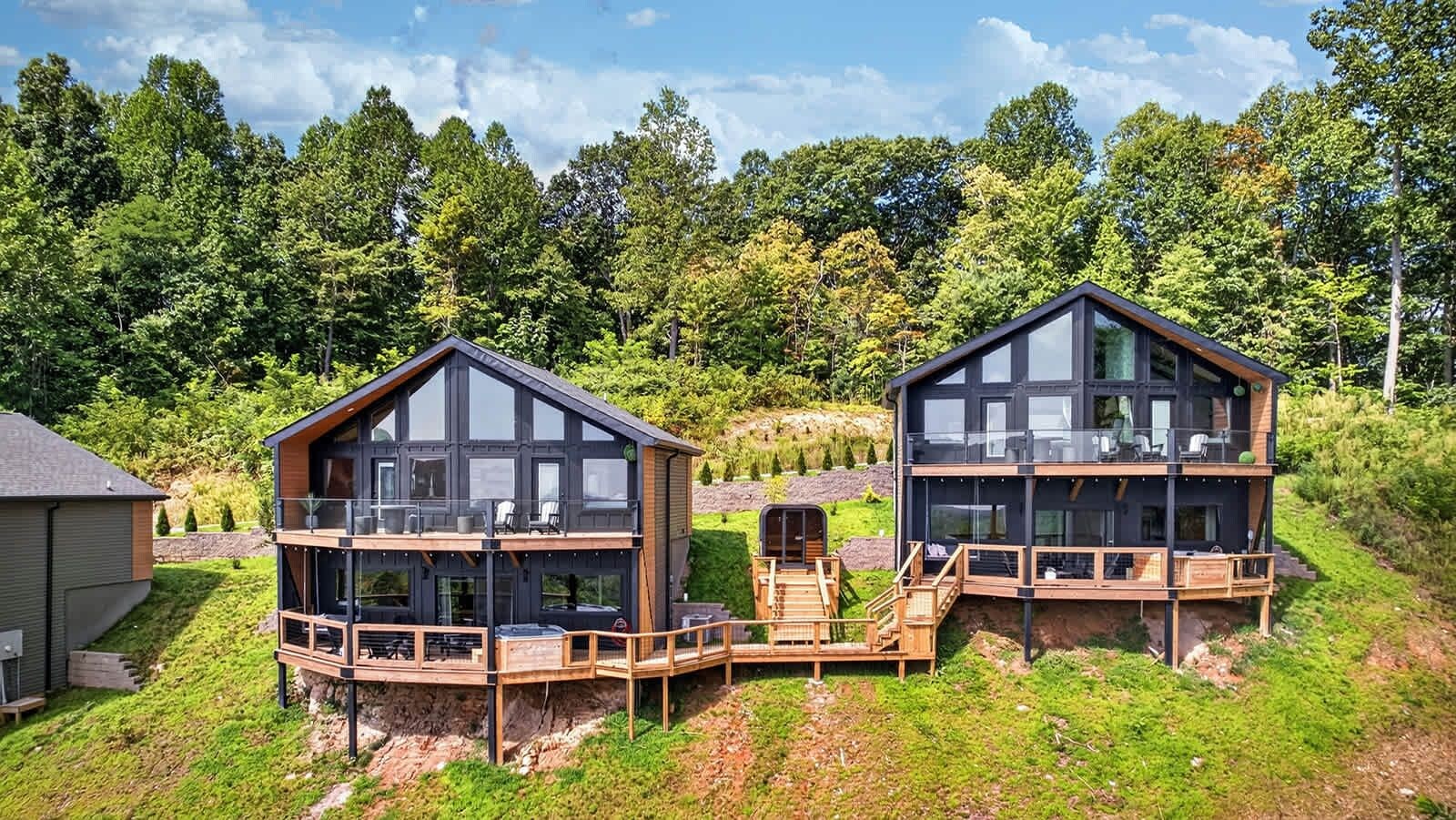 Two modern, multi-story cabins with balconies, situated on a grassy hill with a forest backdrop.