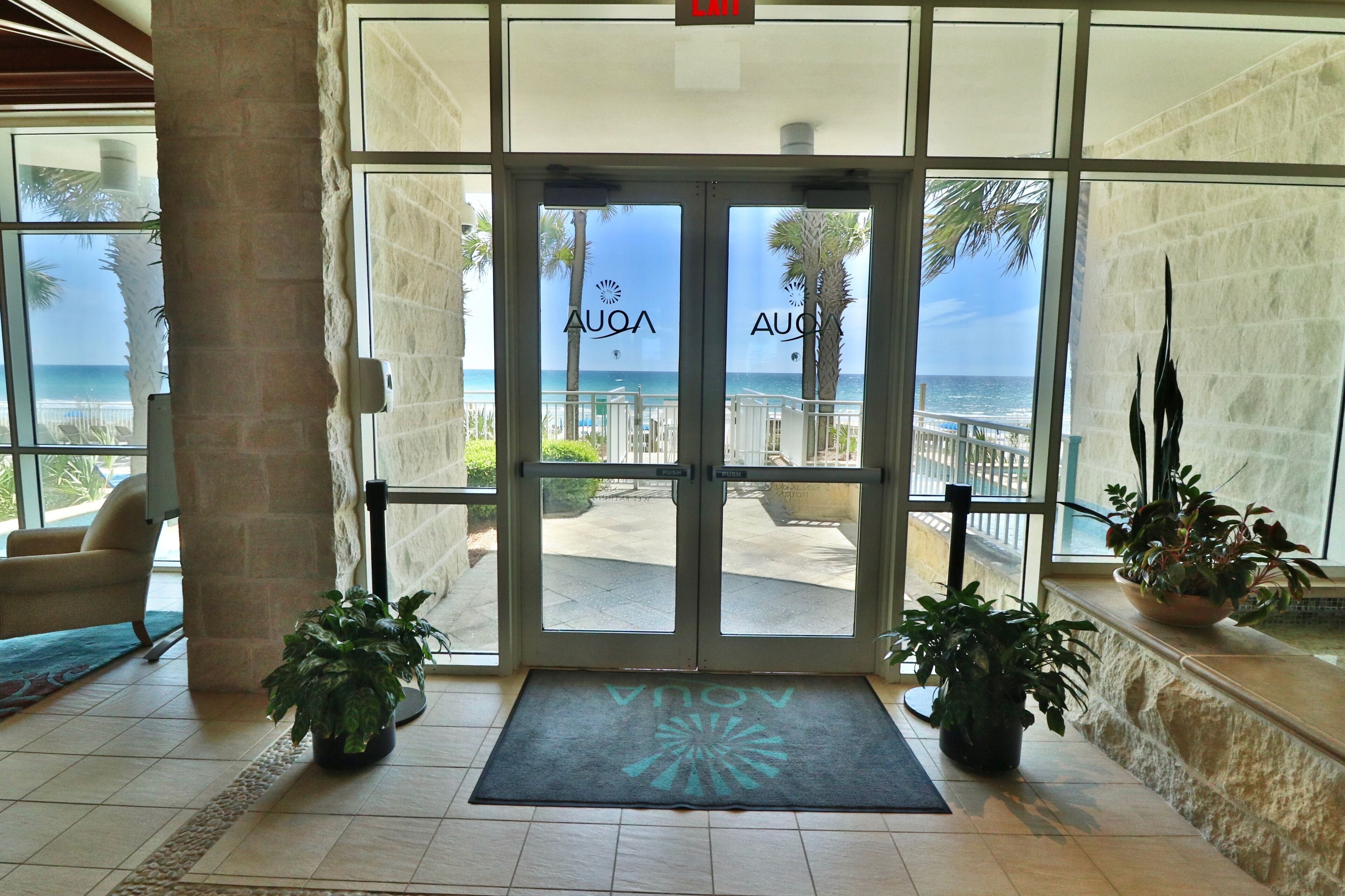 A hotel entrance with a view of the ocean through glass doors, a doormat with the hotel's logo, and potted plants on either side.
