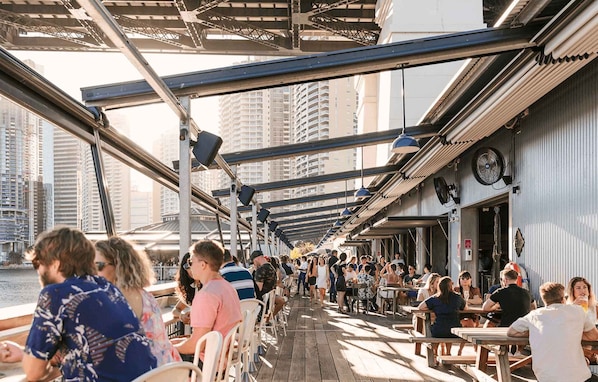 An outdoor dining area with tables and chairs, a view of the water and buildings, and a covered walkway.