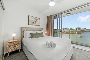 Hotelkamer met een bed, nachtkastje, lamp, schilderij en uitzicht op een rivier en stadsbeeld.